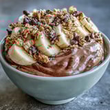 A thick and creamy chocolate peanut butter smoothie bowl garnished with crunchy toppings and a drizzle of maple syrup.